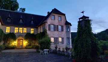 Der Eingang zu Schloss Hochhausen vom Vorplatz aus gesehen. Es ist Abend, die Fenster sind beleuchtet. Rechts vom Schloss steht eine schmale eiserne Stele, auf der ein Krapp zu sehen ist. Die Stele ist umringt von wildem Wein, der auch an der Eingangstür des Schlosses empor rangt.